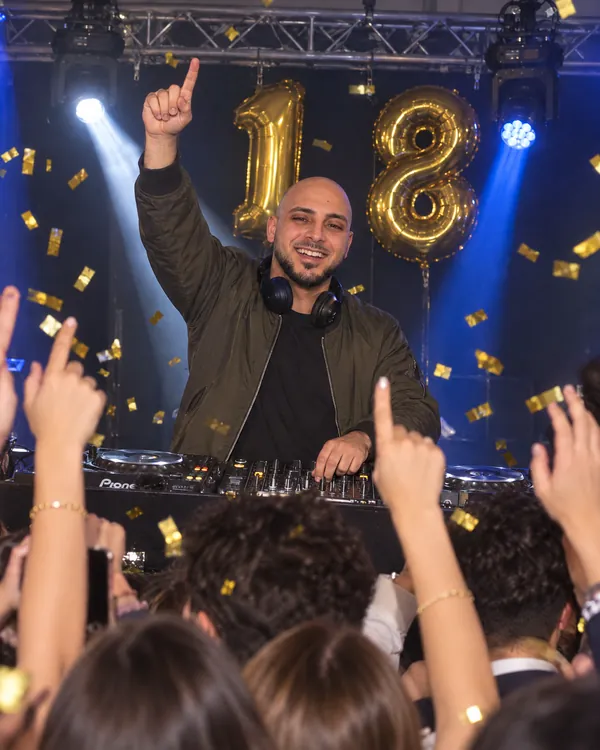 DJ Vlad en bomber kaki, bras levé, ballons 18 dorés et confettis, jeunes qui dansent devant la cabine