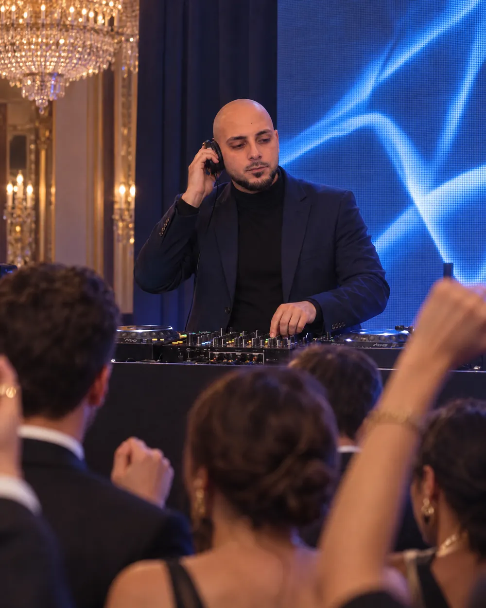 DJ Vlad en blazer marine et col roulé, casque à l'oreille, écran LED bleu derrière lui